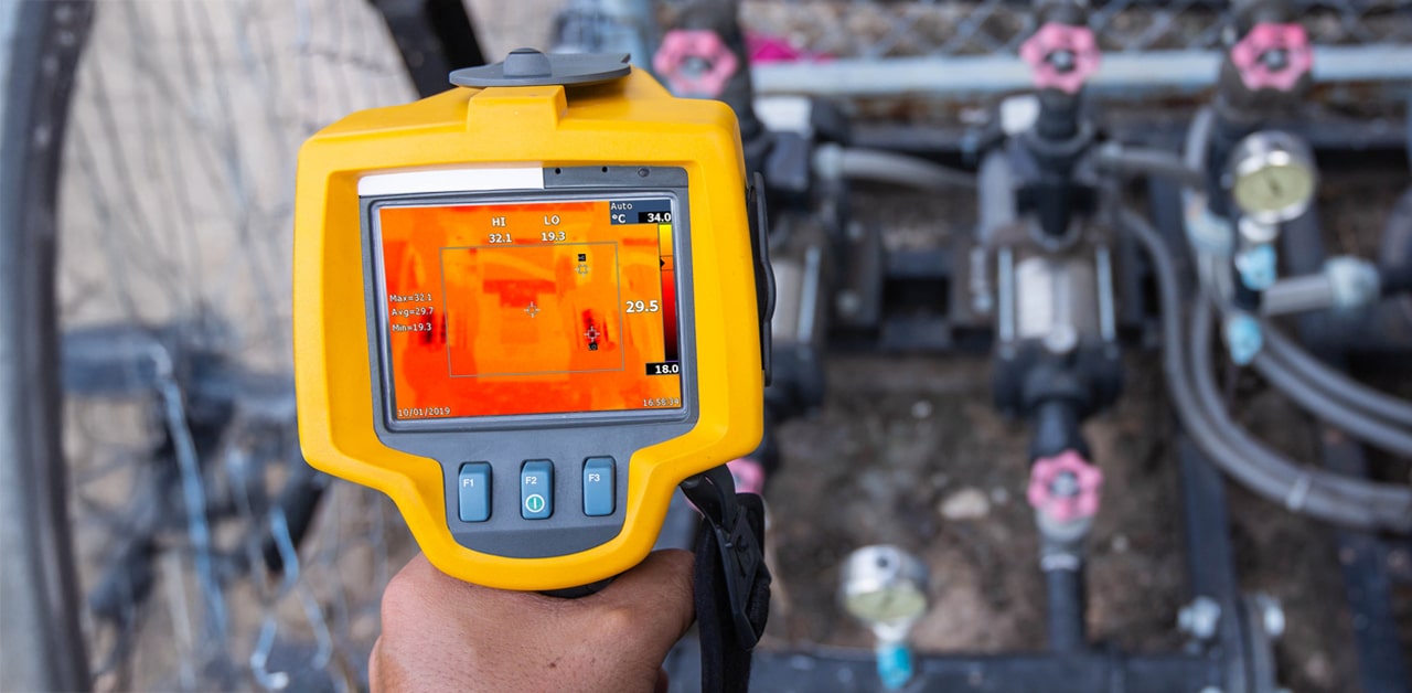 Person holds a thermal camera displaying heat readings of machinery on screen.